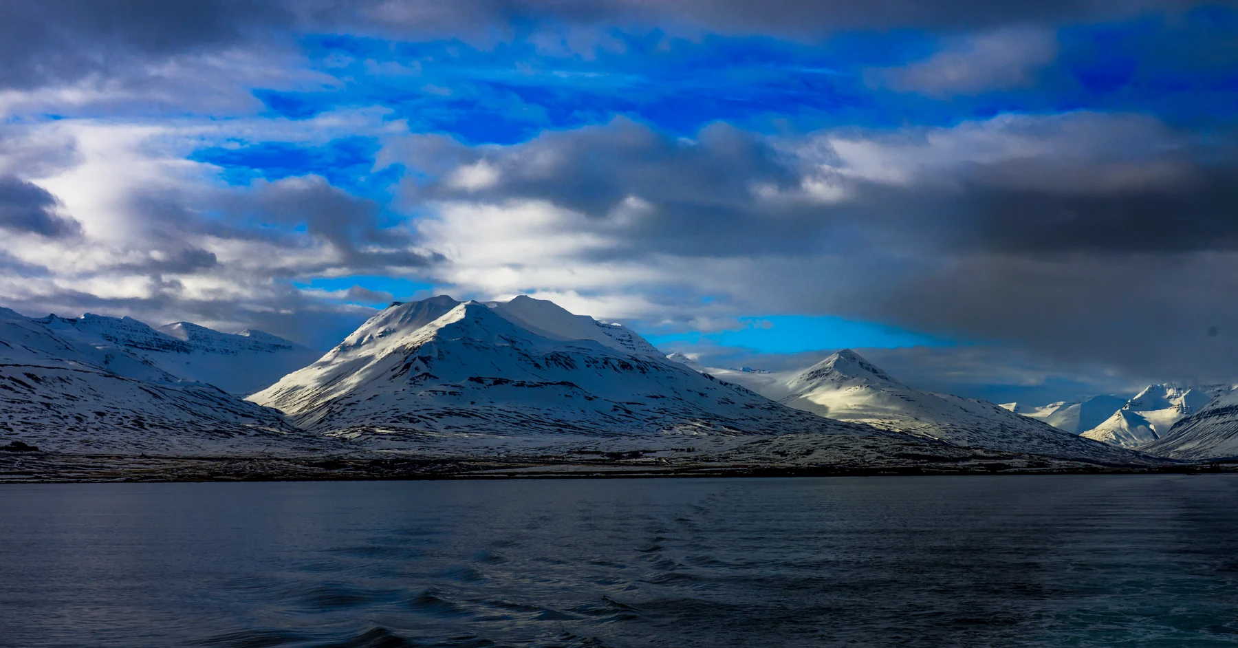 Snow-covered Icelandic mountains across water beneath blue sky and clouds